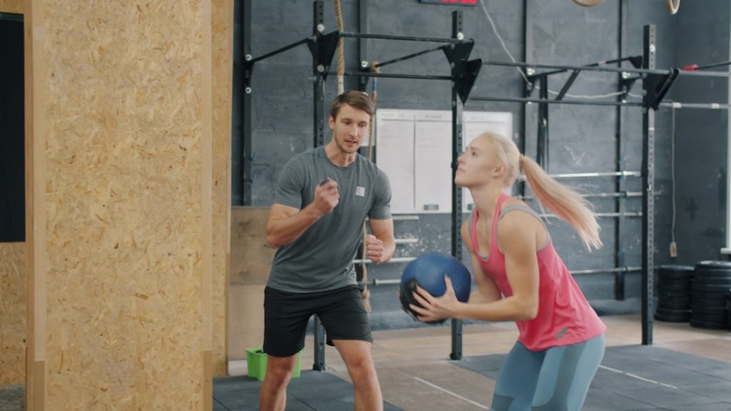 A woman holding a medicine ball with a trainer watching.