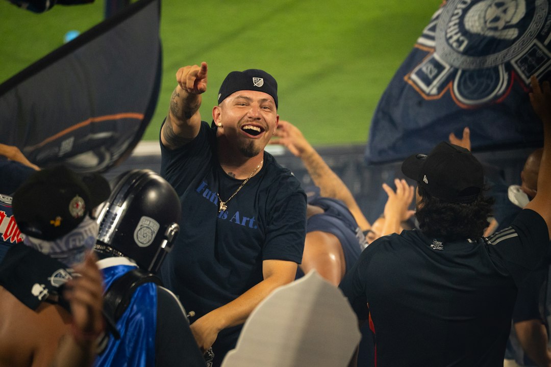 A laughing man in a navy t-shirt pointing at a soccer match